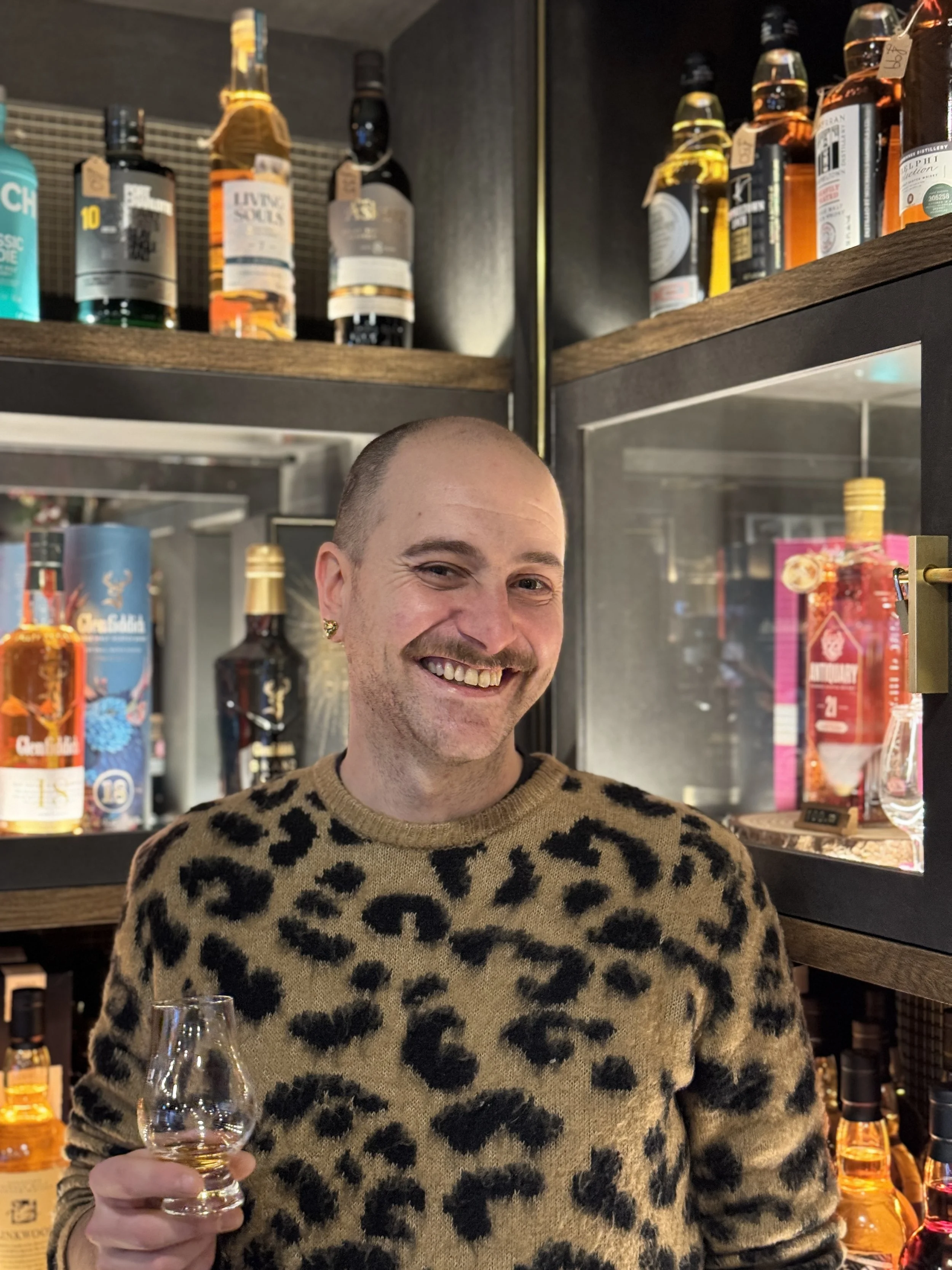 A smiling man with a shaved head and a leopard print sweater holding a glass of whiskey in front of a bar filled with various bottles of alcohol.