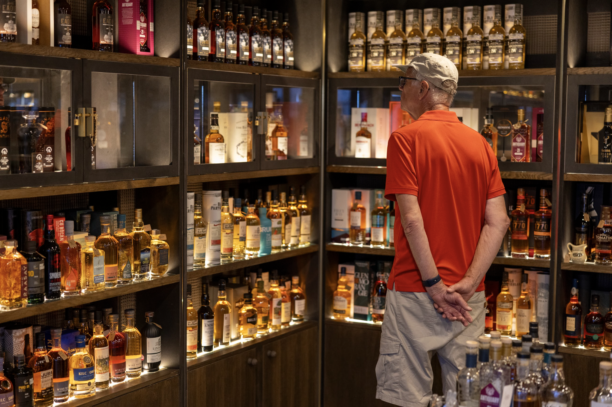 An older man wearing a light-colored cap, glasses, a red polo shirt, and khaki shorts stands in front of a display of whiskey bottles in a liquor store. The shelves behind him are filled with various bottles of liquor, primarily whiskey, in a well-lit retail environment.
