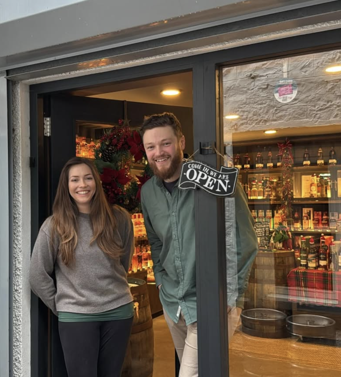 Smiling man and woman standing at the entrance of a store with a sign that says 'Come in, we are open,' and Christmas decorations inside.