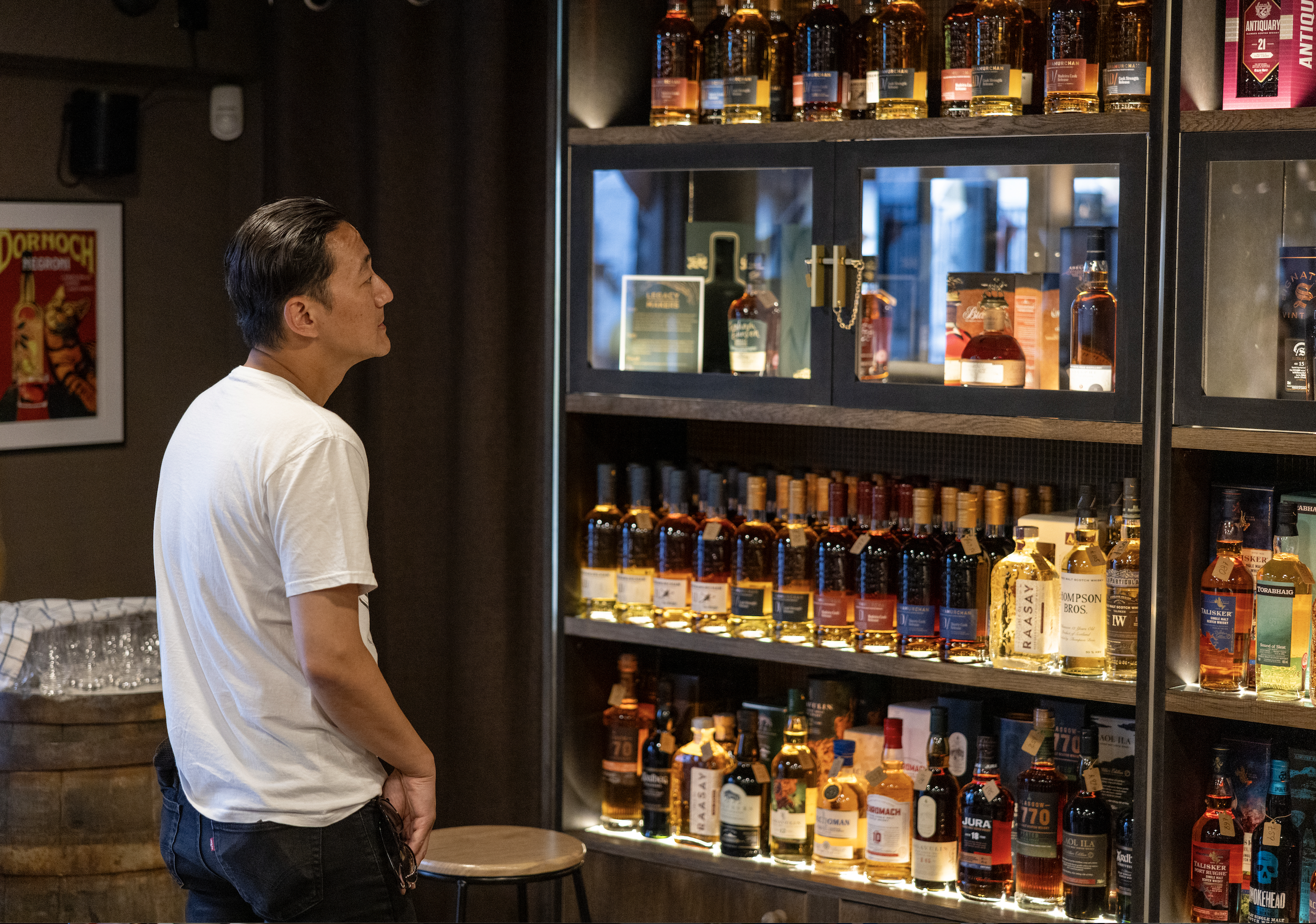A man in a white t-shirt looking at a shelf of various bottles of whiskey or liquor in a whisky store.