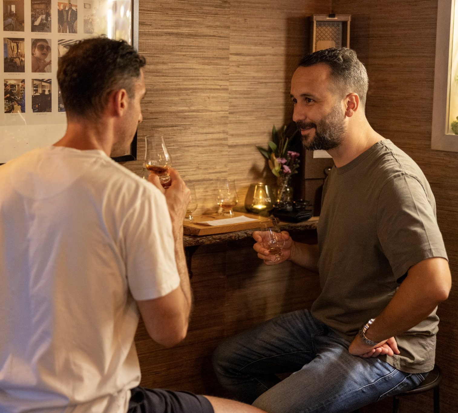 Two men having a conversation in a cozy, warmly lit room, each holding a glass of whiskey, with a photo collage on the wall behind them.