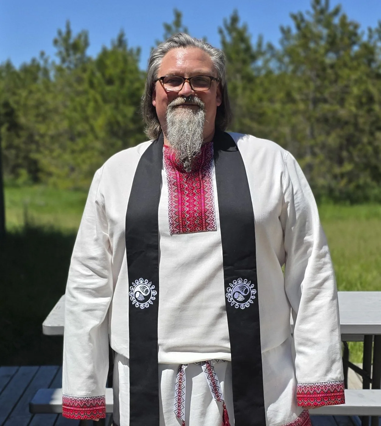 A photo of our Elder, Bear, in white ritual clothing with a black stole. On a wooden deck in front of grass and trees.