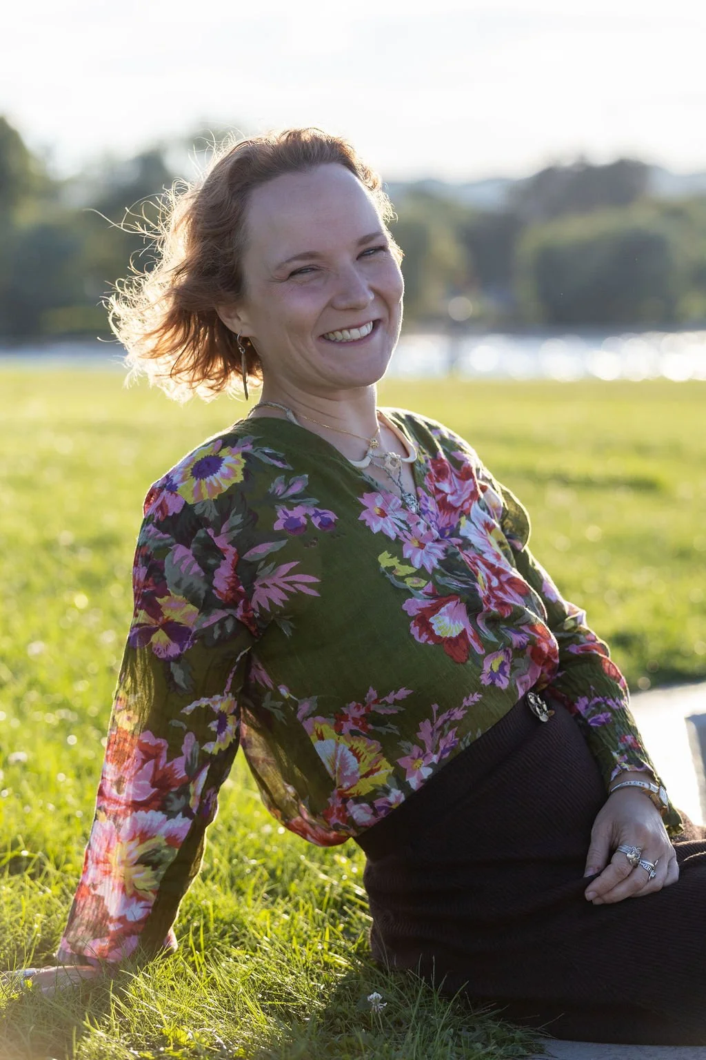 This is an image of Joy sitting on the grass in a green blouse with flowers on it and a big smile with the wind through her hair. The sky is light blue and in the distance you can see trees and the bay
