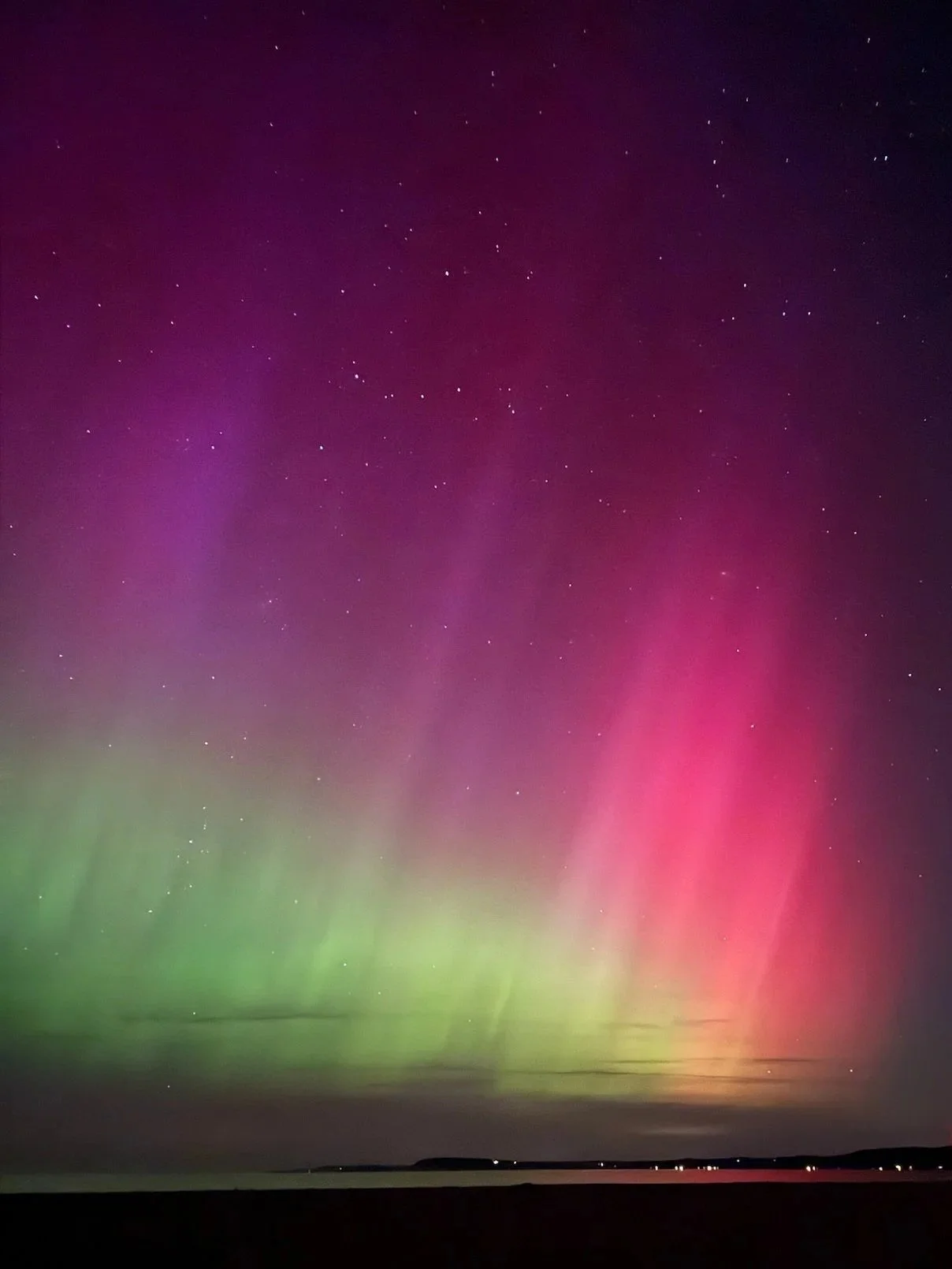A photo of the Aurora Borealis (or Northern Lights.) over the short of Lake Michigan. The sky shows greens, yellows, reds, and purples.  You can see lights from peoples homes across the lake.