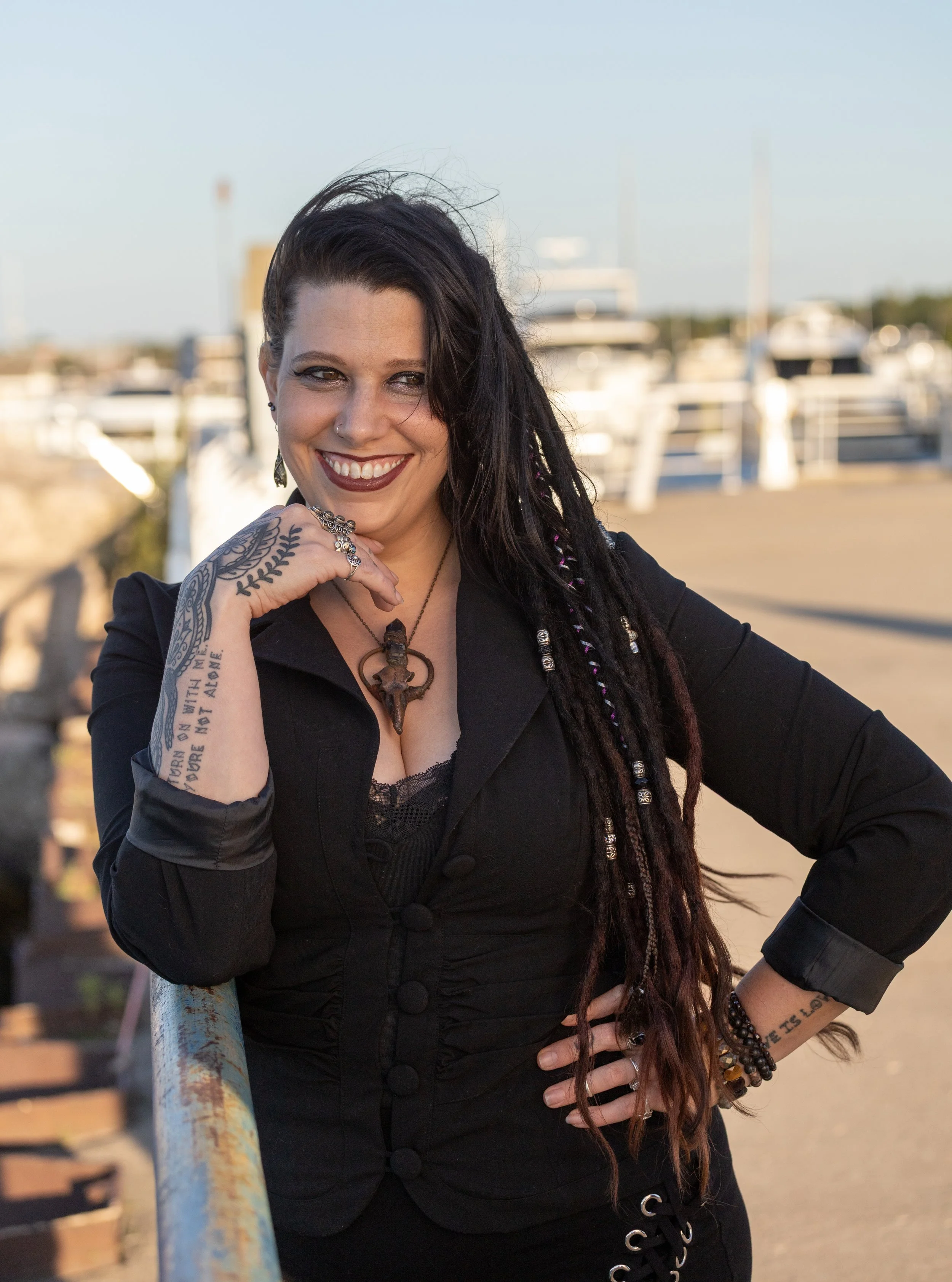 Our own Bellamy Black standing at the edge of Traverse City's Open Space leaning on the railing. She is smiling big, wearing a black outfit with a magickal pendant, and you can see the tattoos on her right hand. There are boats in the background.