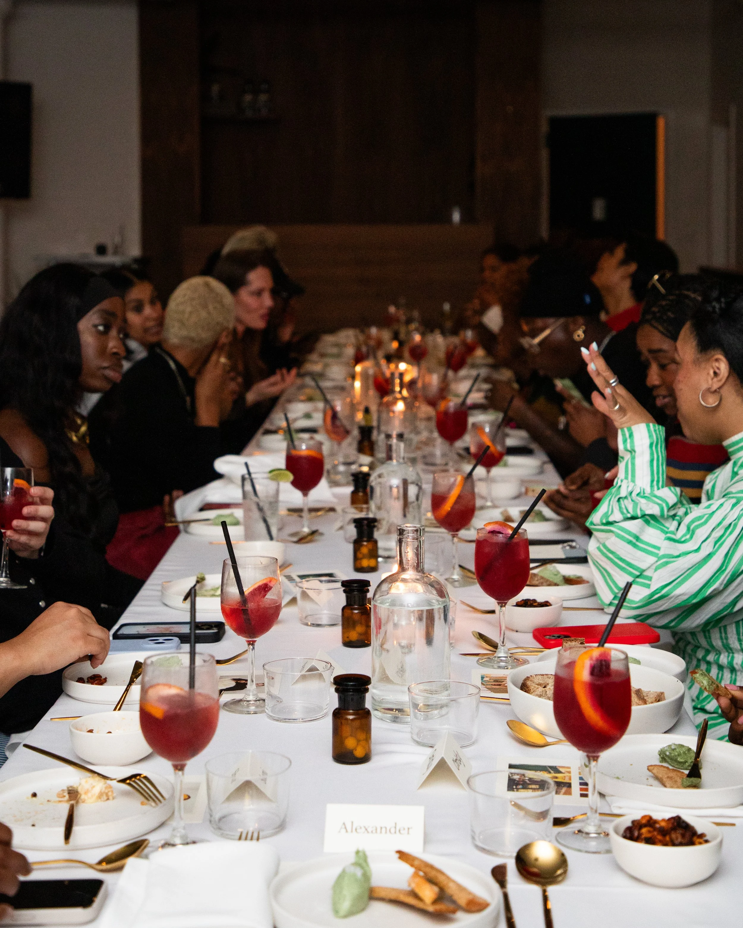 People sitting at a long dinner table during a celebration, with drinks, dishes, and utensils, in a dimly lit room.