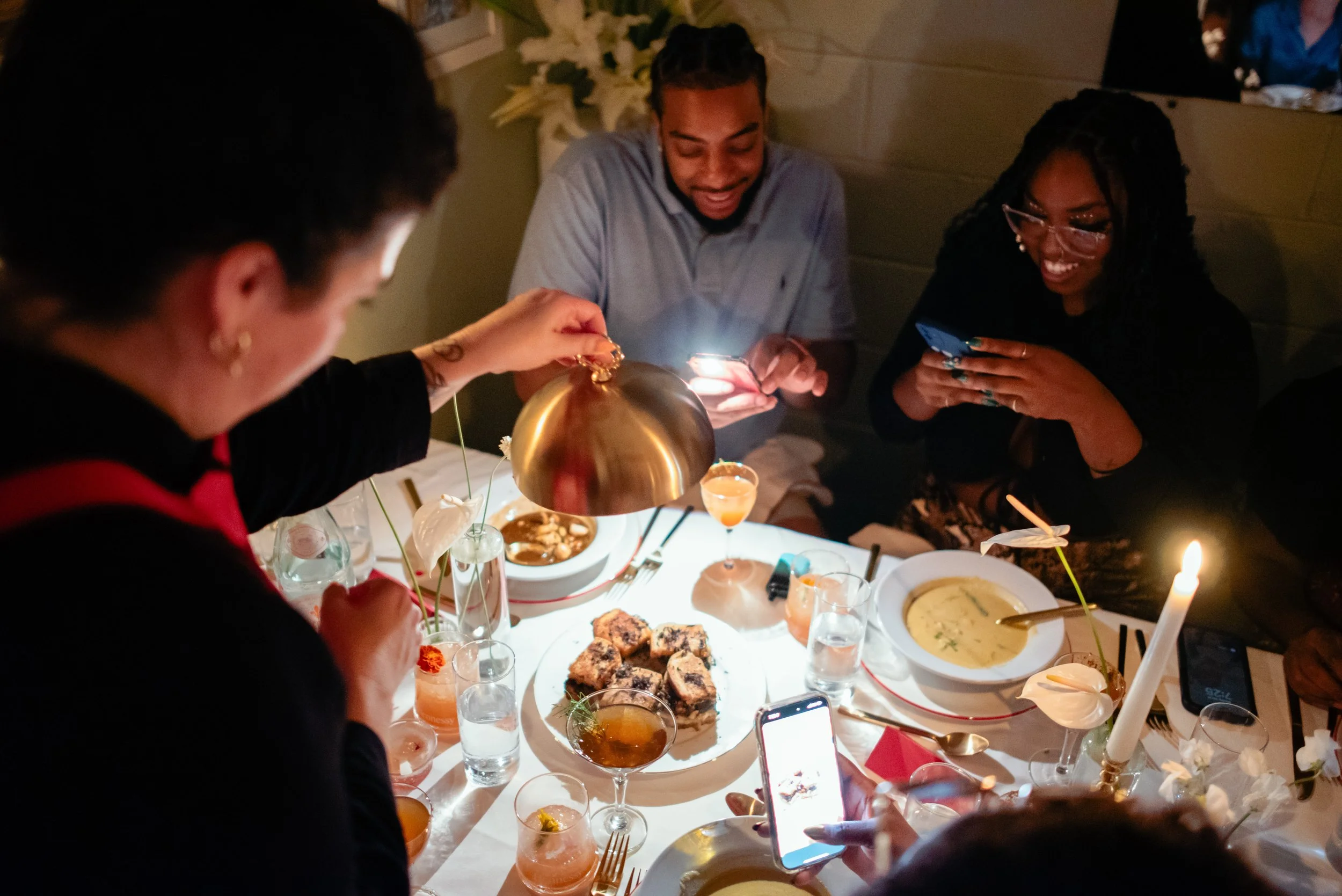People celebrating a brand dinner at a festive table with various foods, drinks, and decorations, including a small lit candle on a cake.