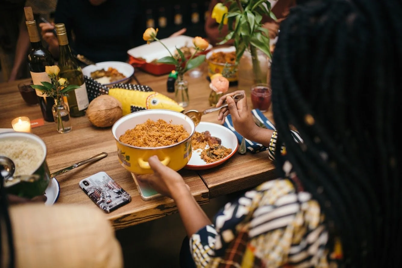 People gathered around a wooden table sharing a meal, with dishes of rice, bowls of food, wine bottles, flowers, and a smartphone on the table.