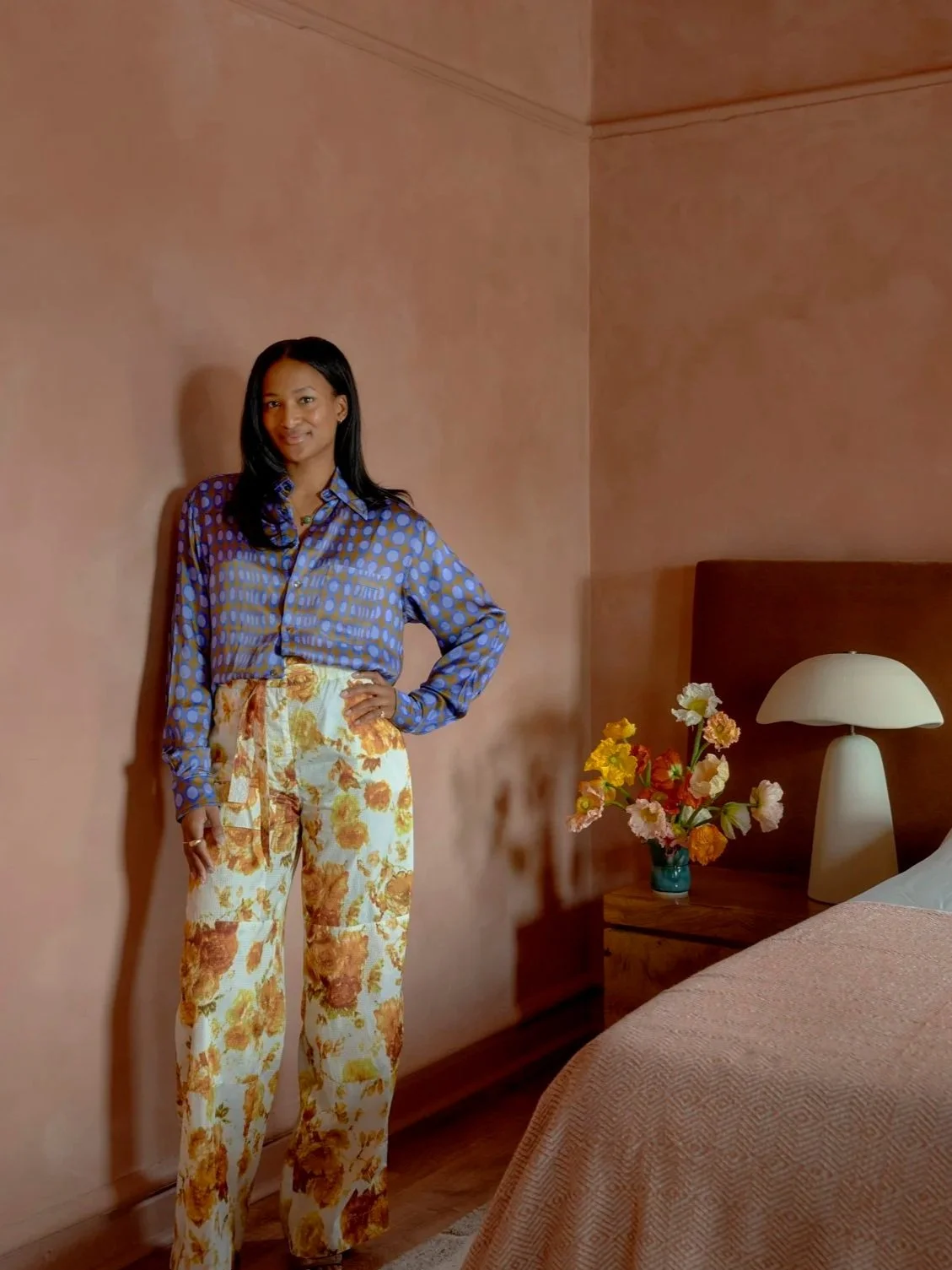 Shanika Hillocks standing in her bedroom, wearing a blue polka dot blouse and floral wide-leg pants, with a vase of flowers and a lamp on a bedside table.
