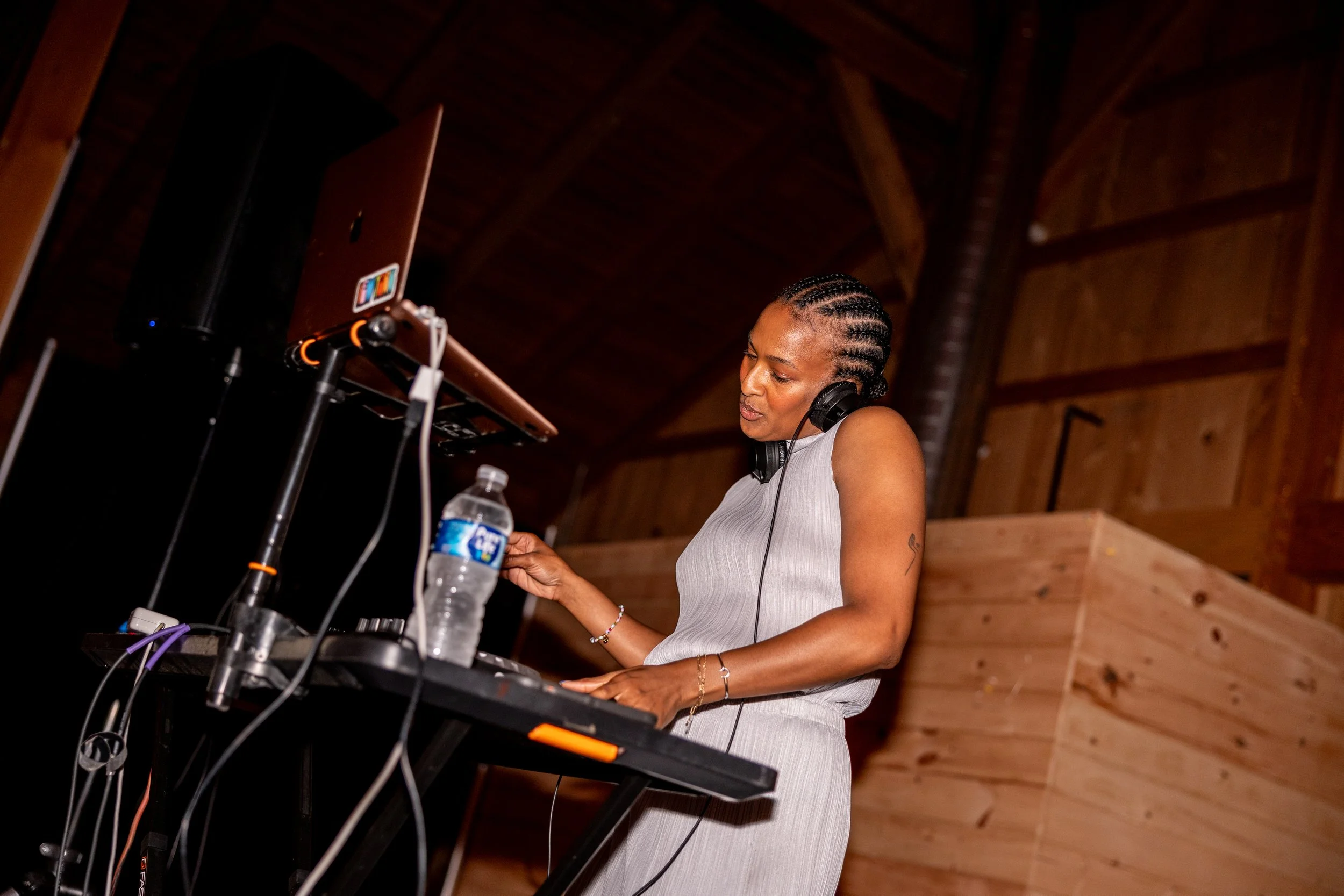 A woman with cornrow braids in a light-colored dress wearing headphones, standing behind DJ equipment with a water bottle on the table in a wooden indoor venue.