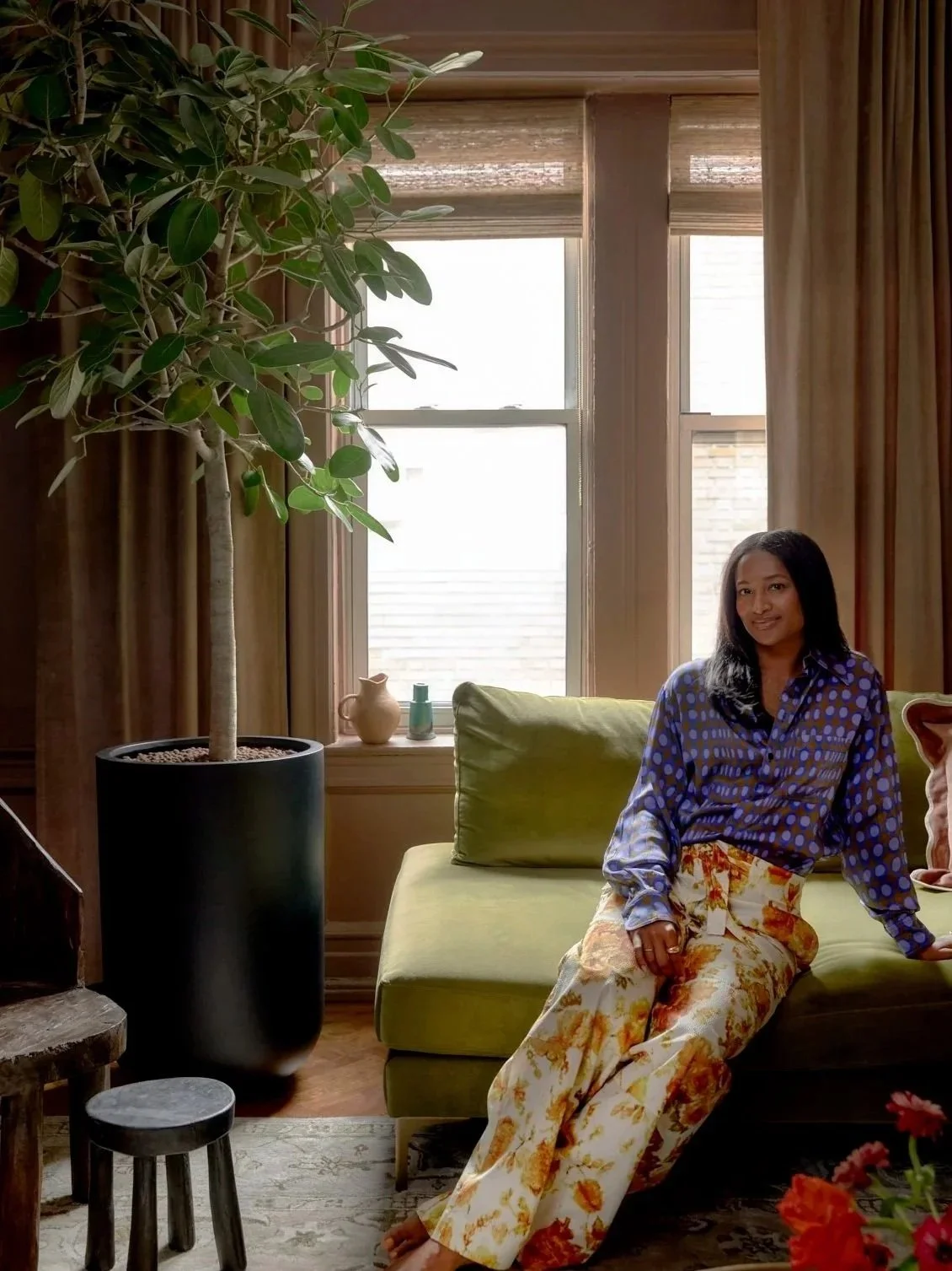 Shanika Hillocks sitting on a green sofa with floral pants and a patterned blue shirt, in a living room with a large potted plant, windows with curtains, and decorative items on the windowsill.