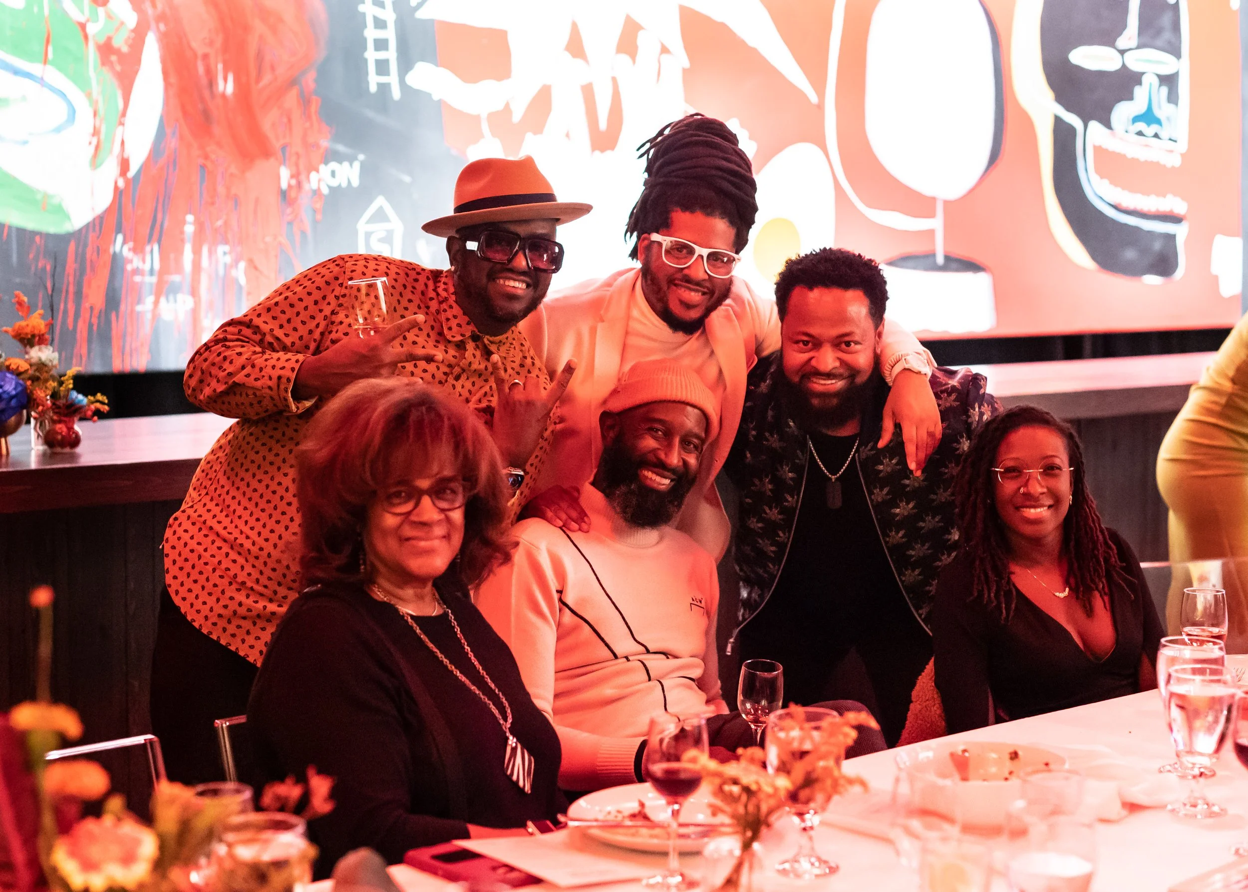Group of people smiling and posing at a dinner party or gathering, sitting and standing around a table with plates and glasses, in a modern decorated room with an abstract mural in the background.