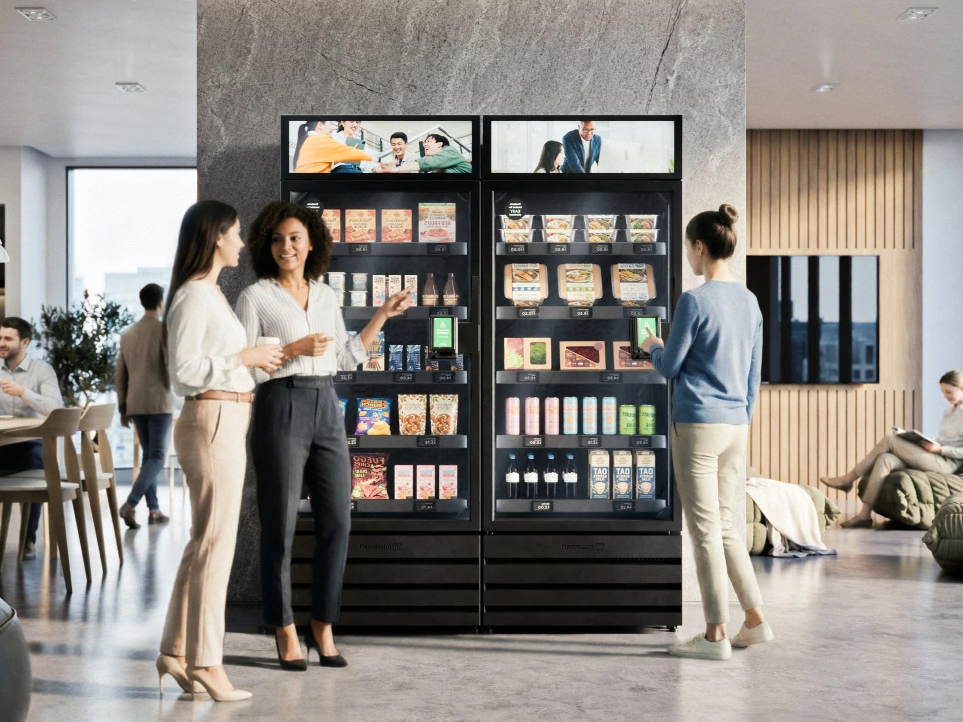 A vending machine in a modern lounge area with three women around it, two of whom are using their phones, and one is talking to the woman on her left. In the background, other people are sitting and standing in the lounge.