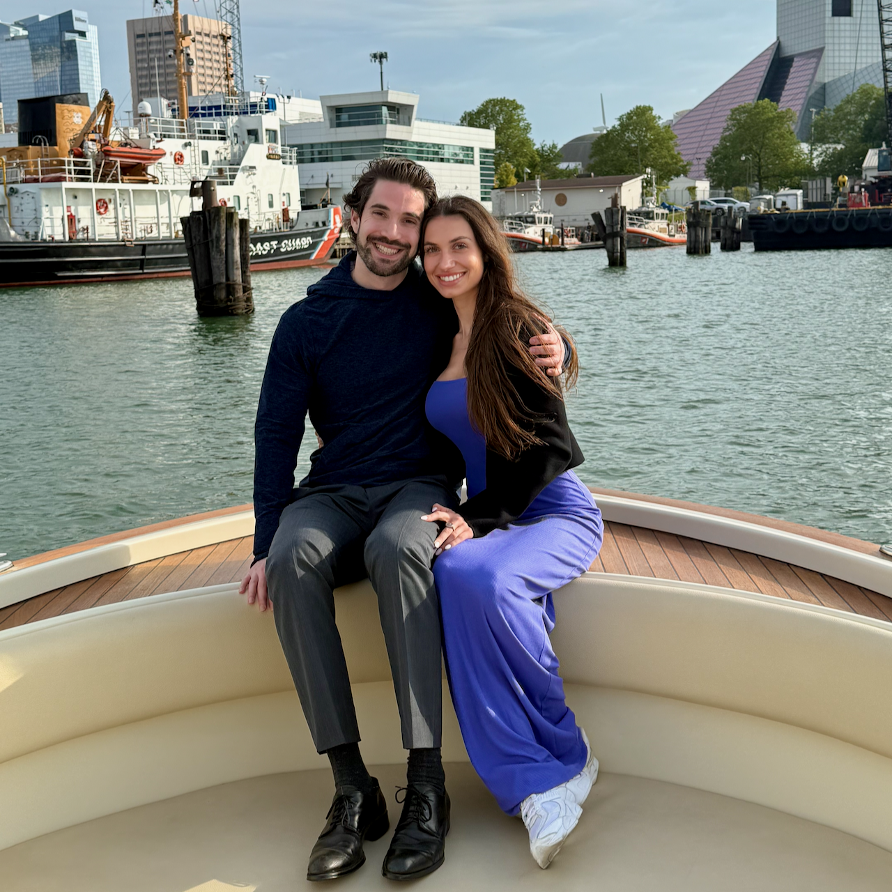 A smiling couple sitting on the edge of a boat, with a city waterfront and several docked boats in the background.