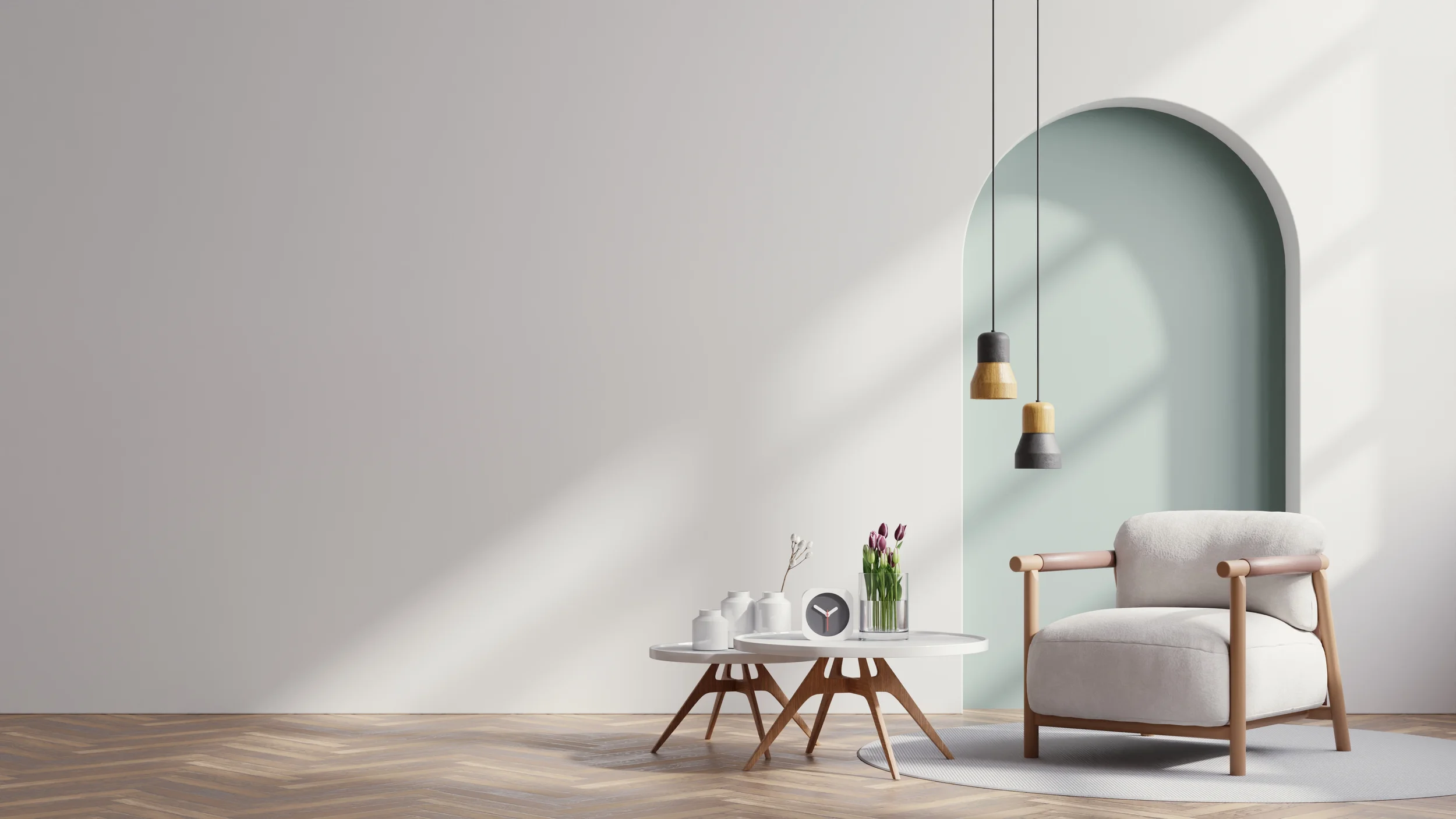 Minimalist living room with a white armchair, a round white coffee table with wooden legs, modern hanging pendant lights, a vase with tulips, a small clock, and candles, against a light blue arched wall and light hardwood flooring.