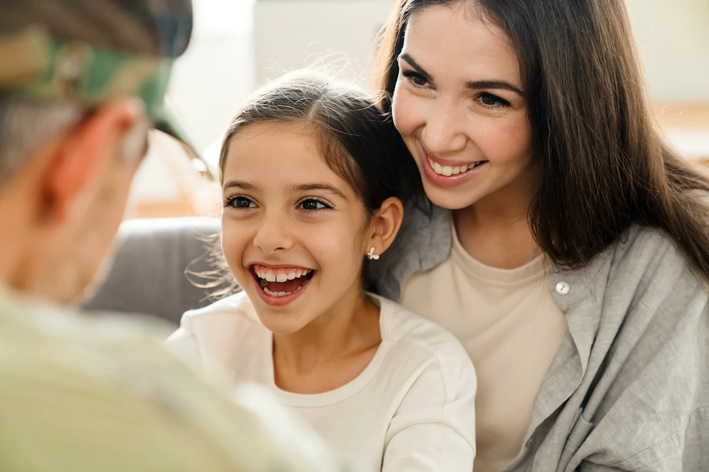 A smiling woman with long brown hair and a young girl with brown hair and earrings, sitting close together and looking at an older person who is out of focus in the foreground.