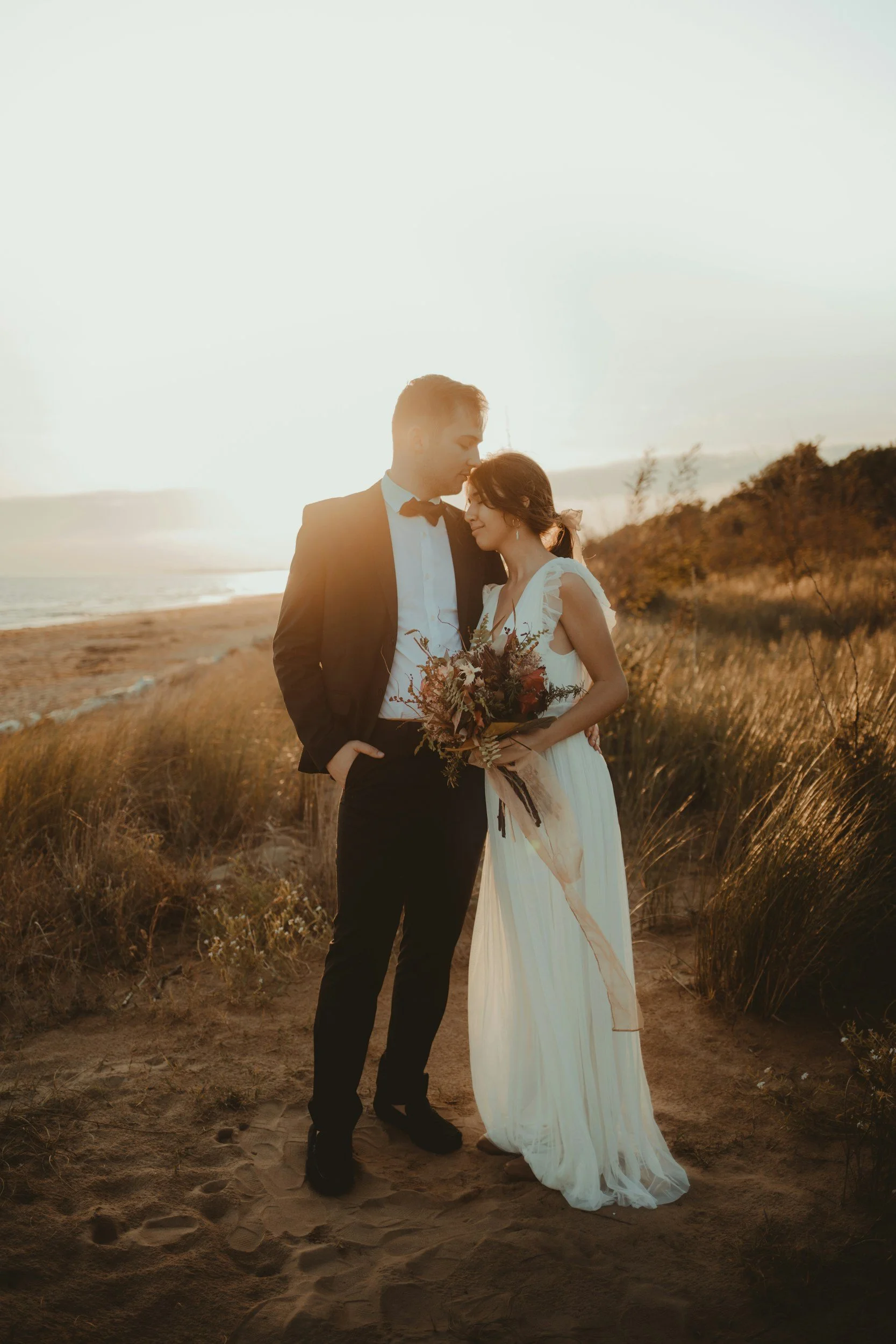 A newlywed couple embraces on a beach at sunset. The groom wears a black tuxedo with a bow tie, and the bride wears a flowing white wedding dress, holding a bouquet of flowers. They stand close together with their foreheads touching, surrounded by grassy dunes and the ocean in the background.