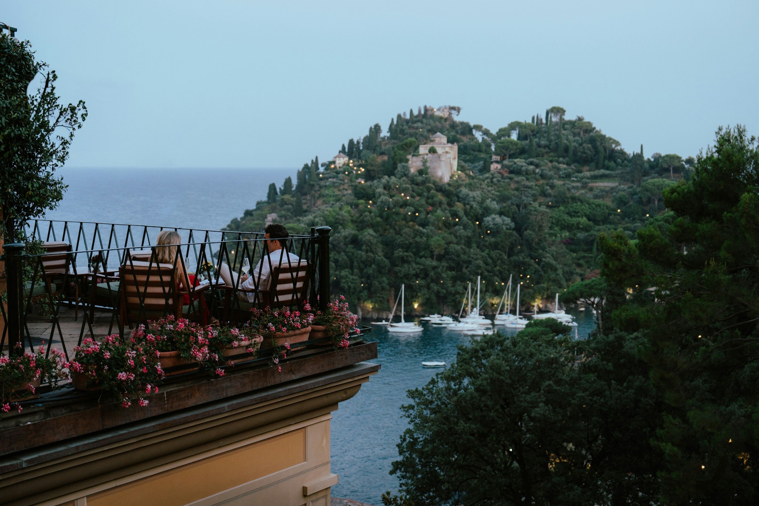 Couple sitting on a balcony with pink flowers, overlooking a harbor with sailboats and a green hillside with trees and buildings.