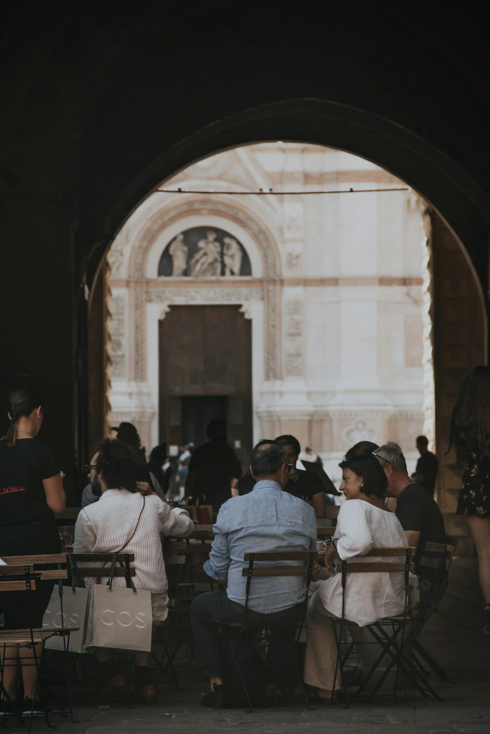 People sitting at tables inside a historic building with ornate architectural details, seen through a large arched window.