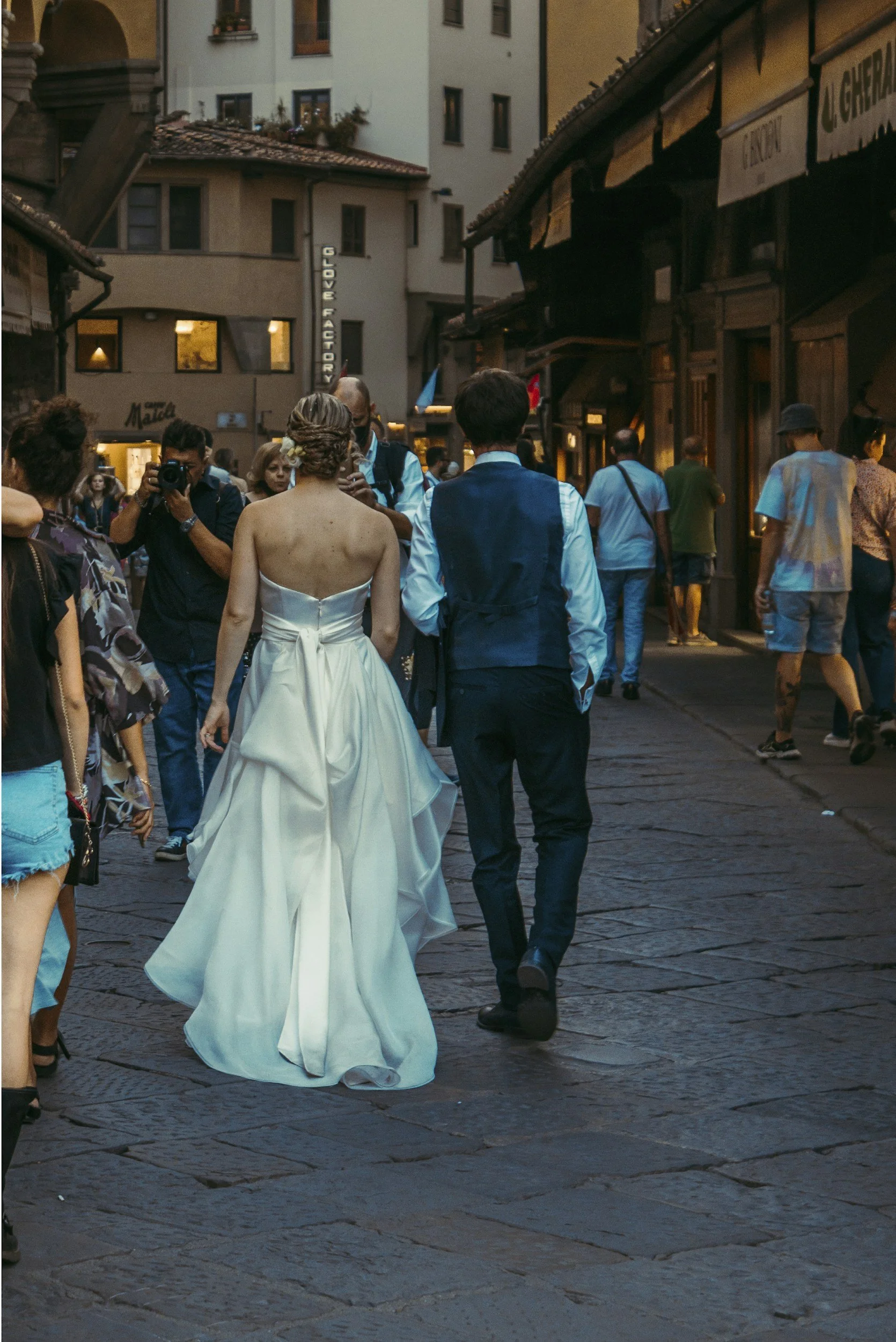 A woman in a white dress and a man in a blue vest and white shirt walking together on a street crowded with people, some taking photos. The background shows buildings with shop signs and windows.