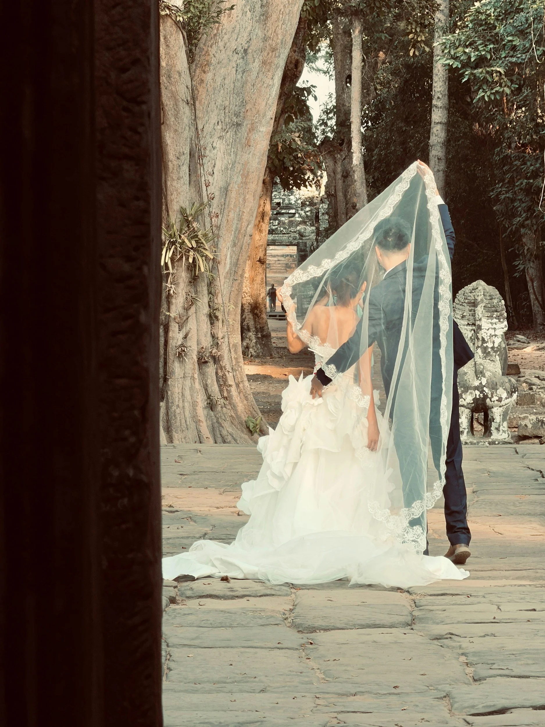 A bride in a white wedding gown and veil, and a groom in a suit, walking outdoors in a historic, wooded area with ancient stone ruins and statues.