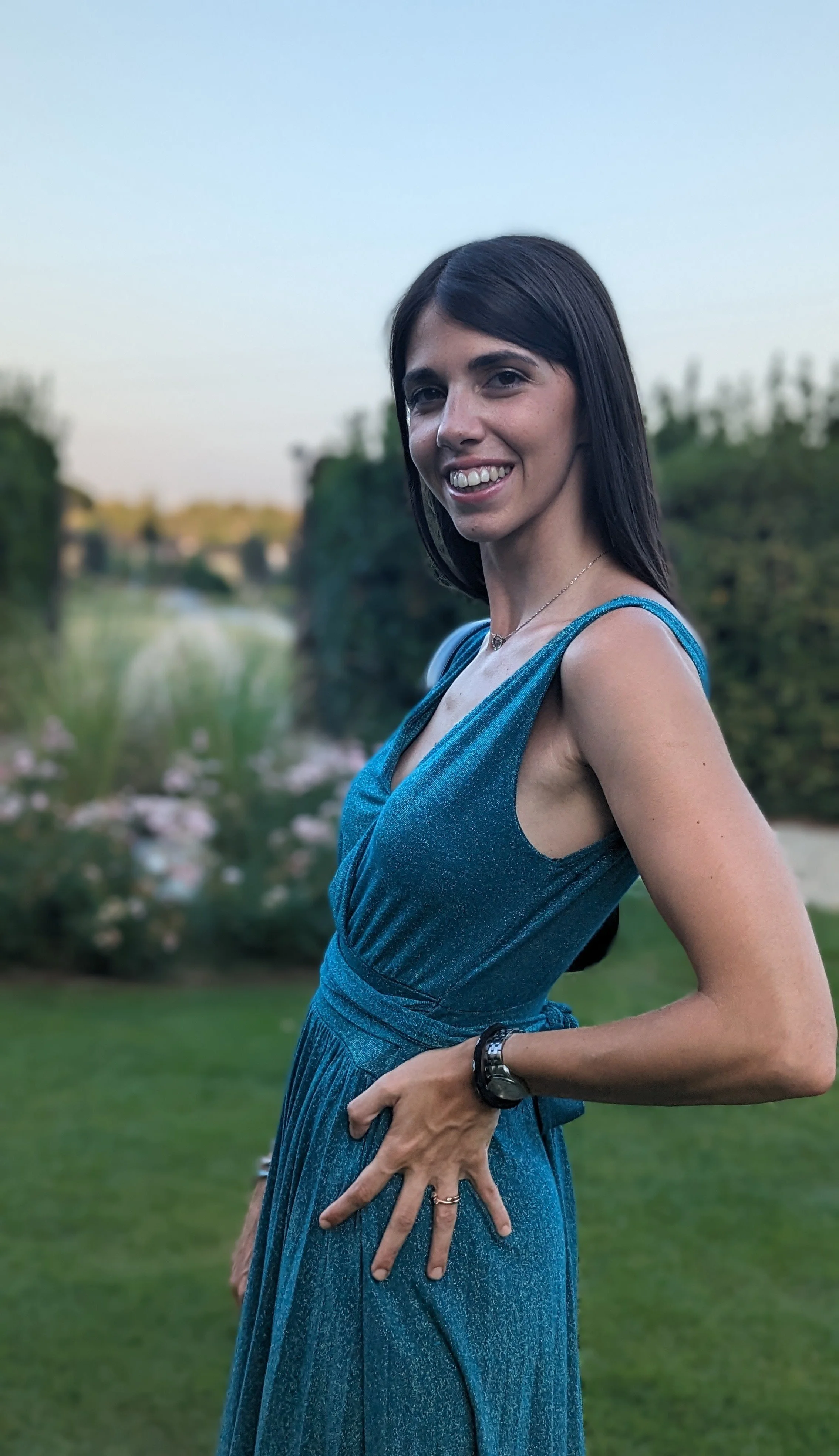 A woman in a shiny blue dress standing outdoors with a blurred green background of trees and water, smiling at the camera.