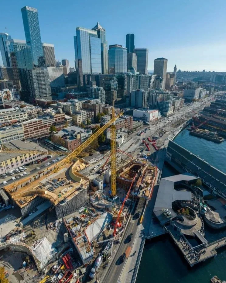Aerial view of a downtown city skyline with skyscrapers, construction cranes, and water surrounding a construction site near the waterfront.