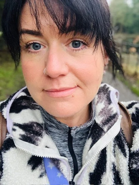 Close-up of a woman with short black hair, light skin, blue eyes, wearing a nose ring, and a black-and-white patterned fleece jacket outdoors.