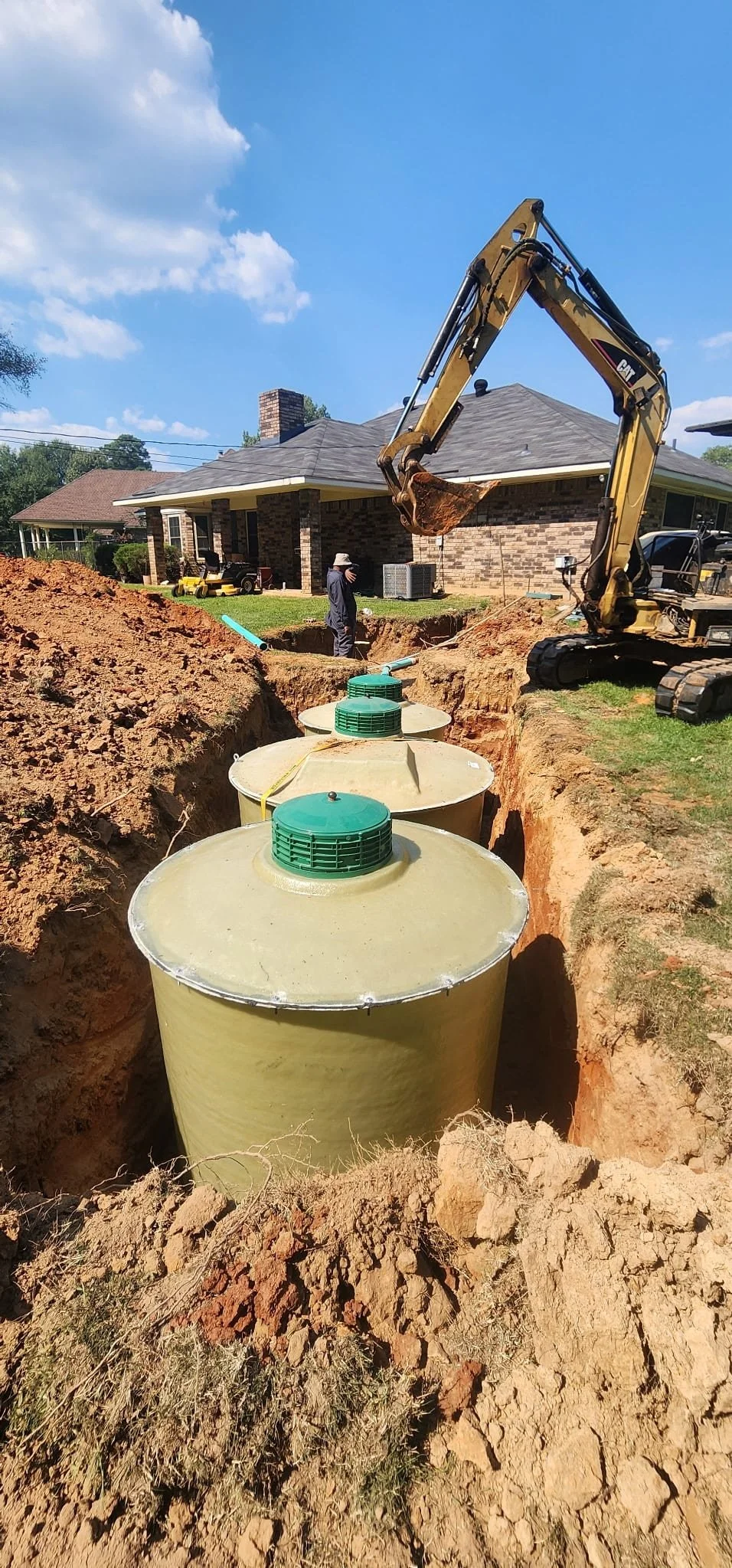 Construction site with underground septic tanks being installed in a yard, with an excavator and workers present.