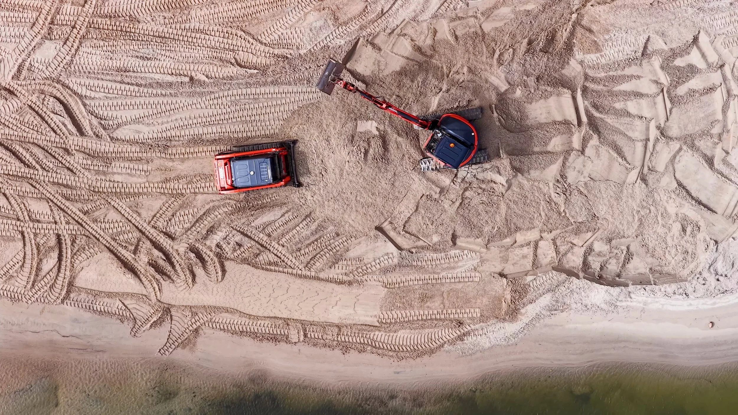 Aerial view of construction equipment and tire tracks in sand or dirt, with a small excavator moving soil.