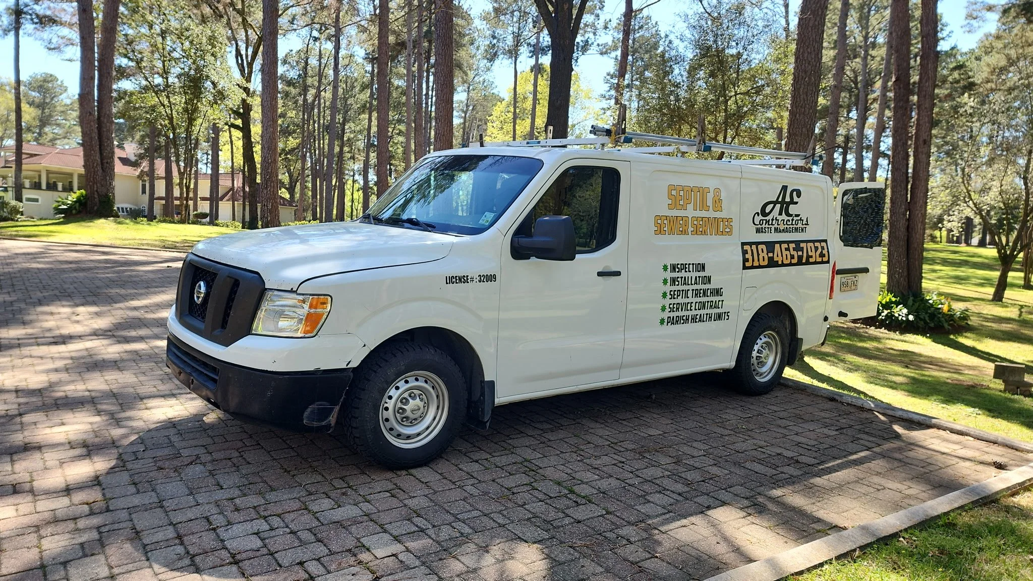White service van parked on a brick driveway in a wooded residential area, advertising septic and sewer services with contact phone number.