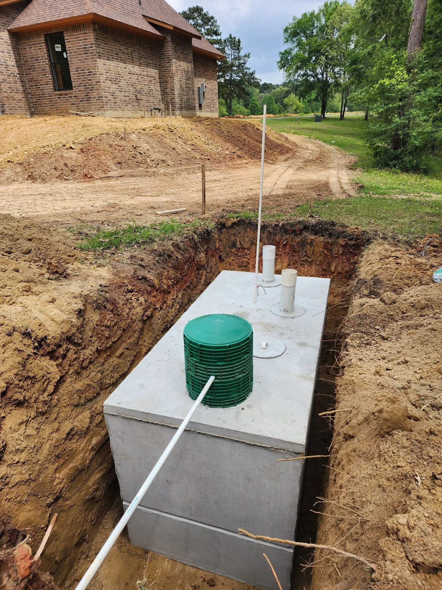 Underground utility installation at a construction site near a brick house, with dirt and holes in the ground, piping, and a green cylindrical water utility structure.