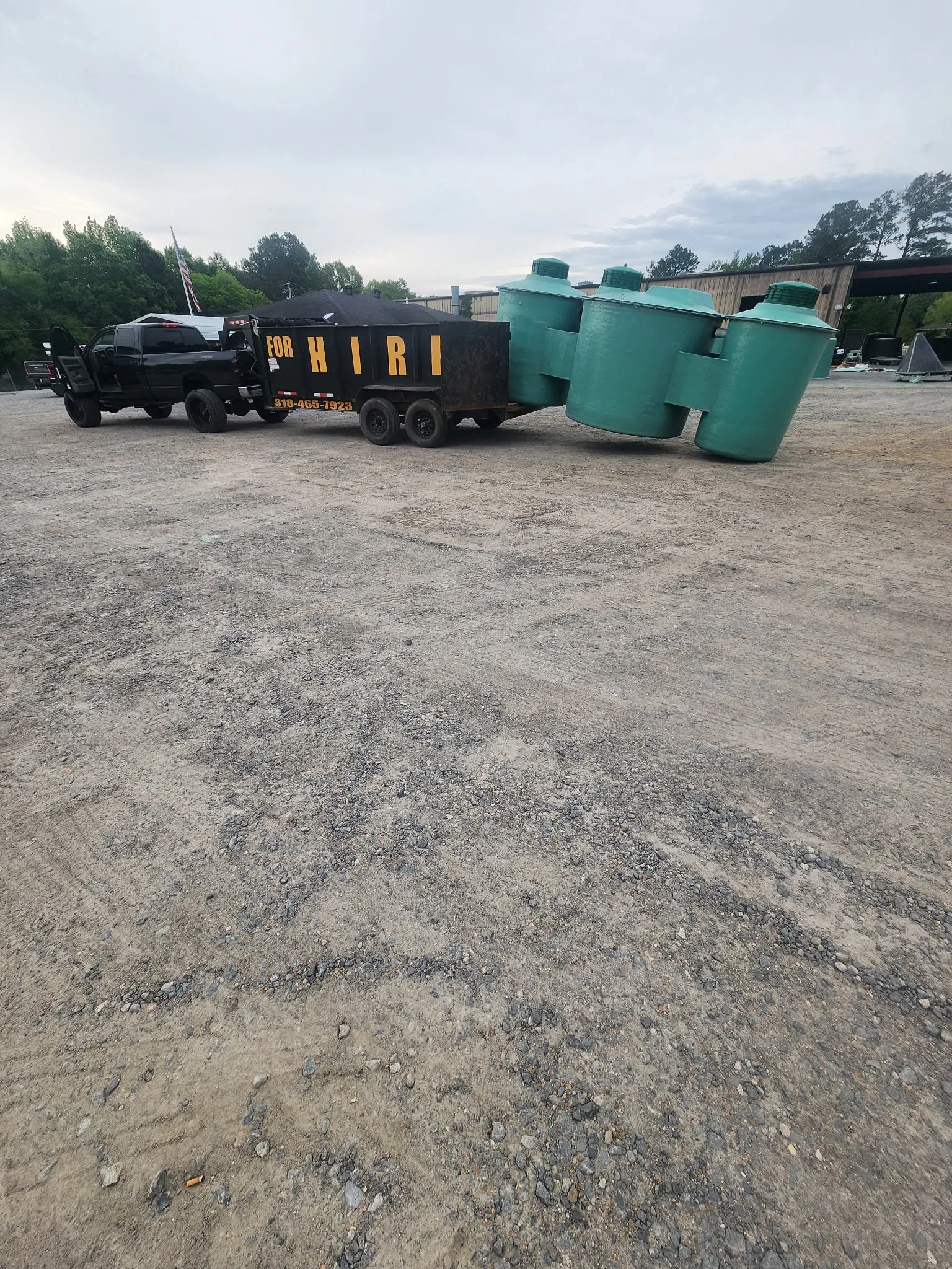 A black truck with a fenced trailer carrying three large green tanks in a gravel lot with a building and trees in the background.