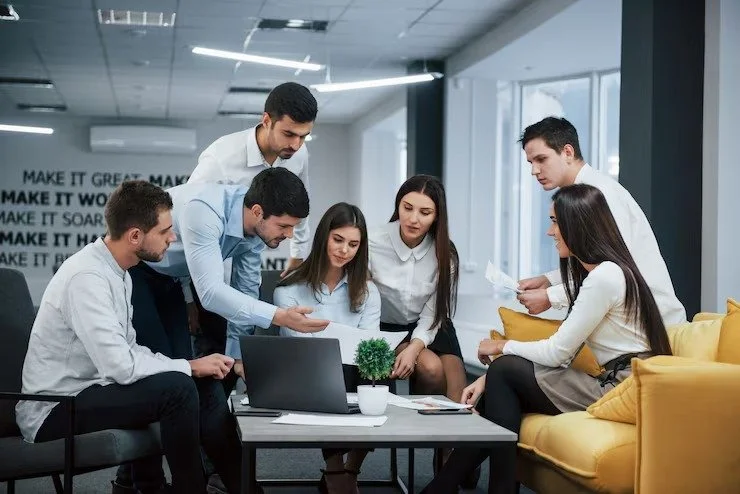 Six young professionals in a meeting room, gathered around a laptop on a coffee table, discussing documents and working together.