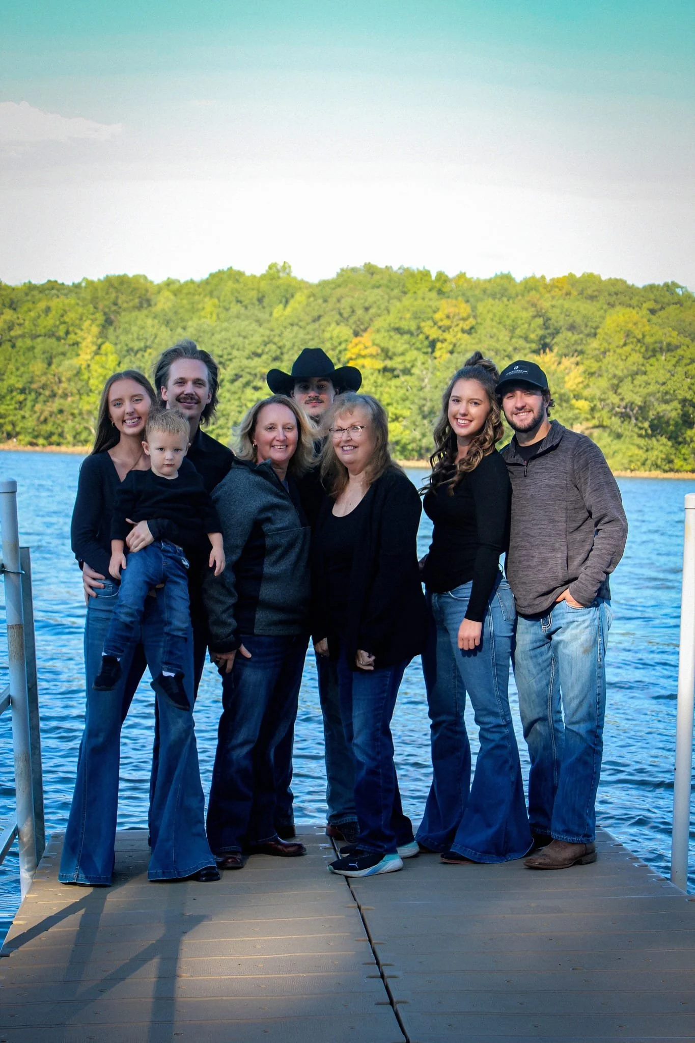 Group of seven people, including a child, standing on a dock by a lake with a forested shoreline in the background, during late afternoon or early evening.