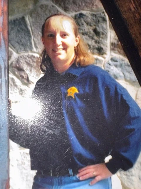 A person with long hair wearing a blue shirt with a badge on the chest, standing outdoors in front of a stone wall.