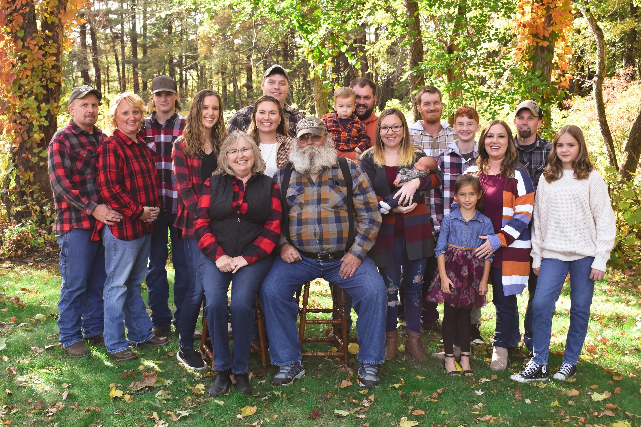 A large multigenerational family portrait outdoors in a forest during autumn, with trees and fallen leaves in the background. The family is smiling, wearing casual fall clothing, and positioned closely together, with some standing and some sitting, including children, adults, and seniors.