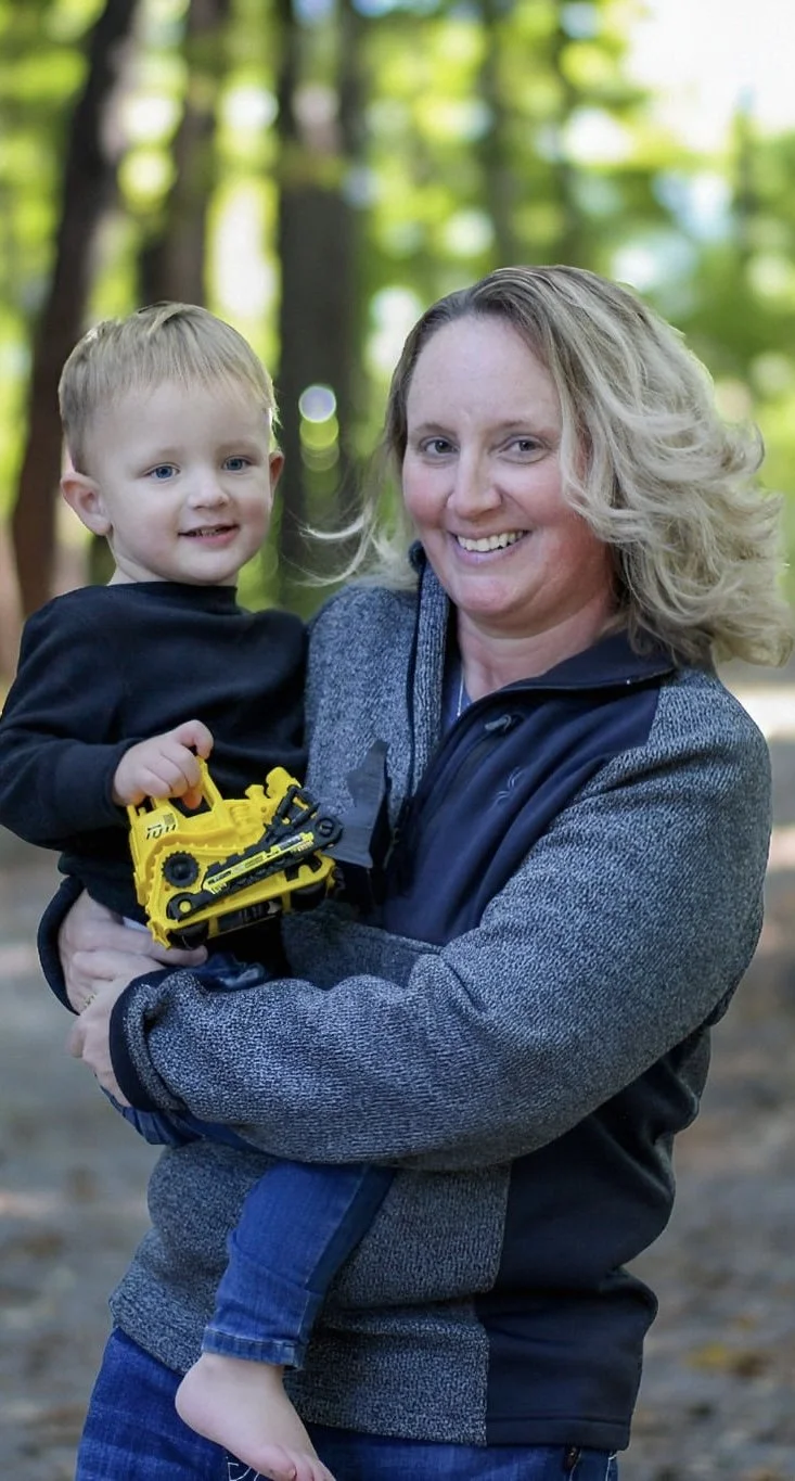 A woman smiling while holding a young boy in an outdoor wooded area.