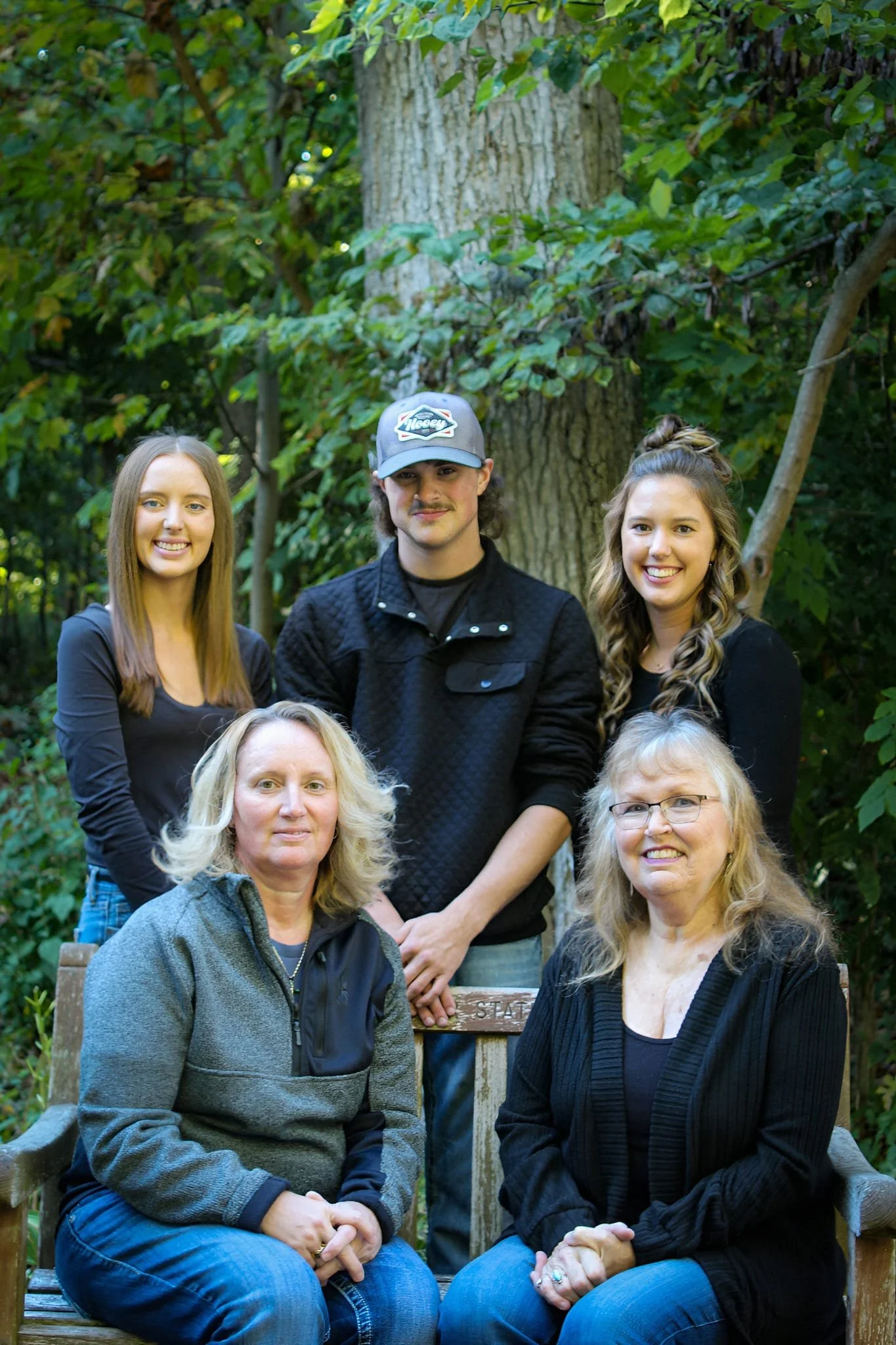 A group of seven people, five women and two men, gathered outdoors in front of a tree and dense greenery, smiling at the camera.