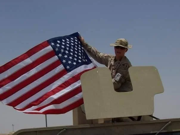 A woman in military uniform holding an American flag from a vehicle window.