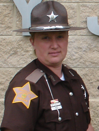 A female law enforcement officer in uniform, wearing a brown hat with a silver badge, a brown shirt with a sheriff's badge on the sleeve, and standing in front of a beige brick wall.
