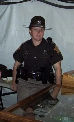 A female police officer in uniform standing behind a table, wearing a wide-brimmed hat, with a badge and equipment on her belt.
