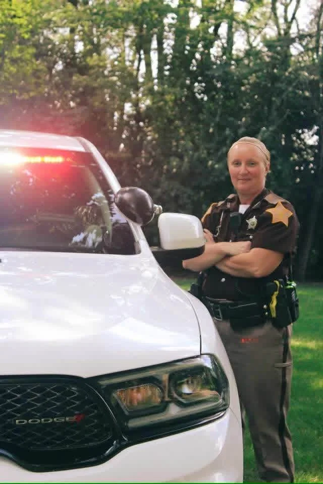 A female police officer standing next to a white police vehicle, with her arms crossed and a slight smile, outdoors with trees in the background.