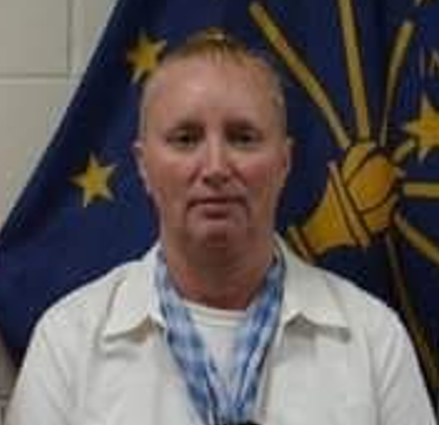 A woman with short light brown hair wearing a white shirt and blue scarf standing in front of a flag with stars and a torch symbol.