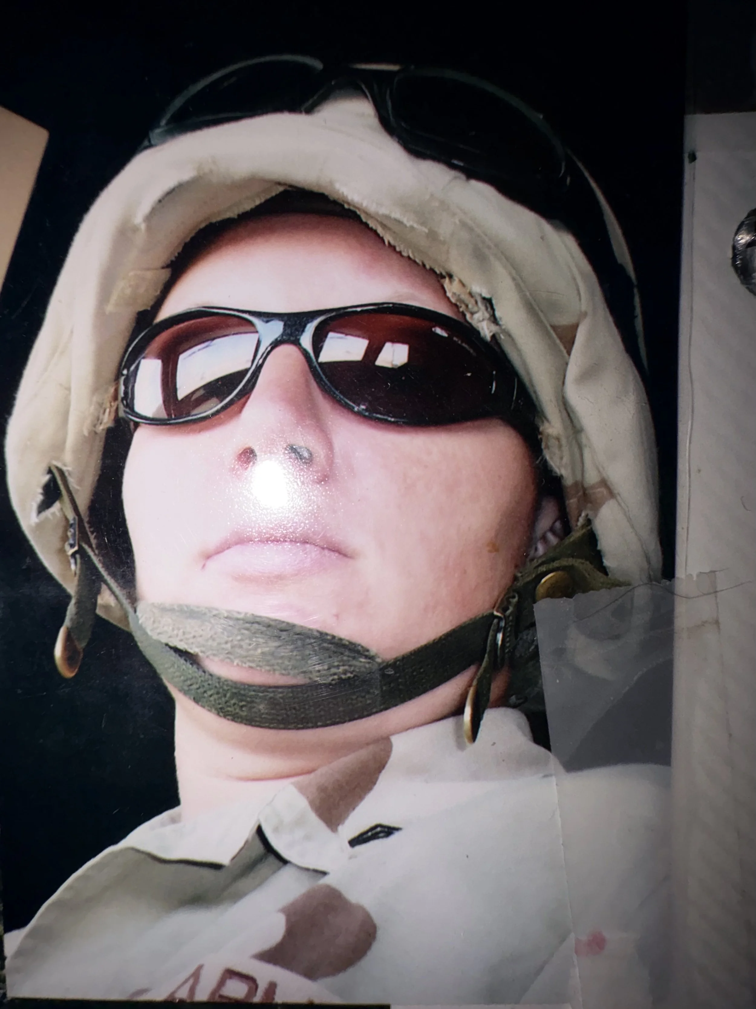 Close-up photograph of a person wearing a military-style helmet and sunglasses, with a white hoodie and a white shirt. The person's face is partially visible, and the photo appears to be taken from above, showing the person's nose and mouth.