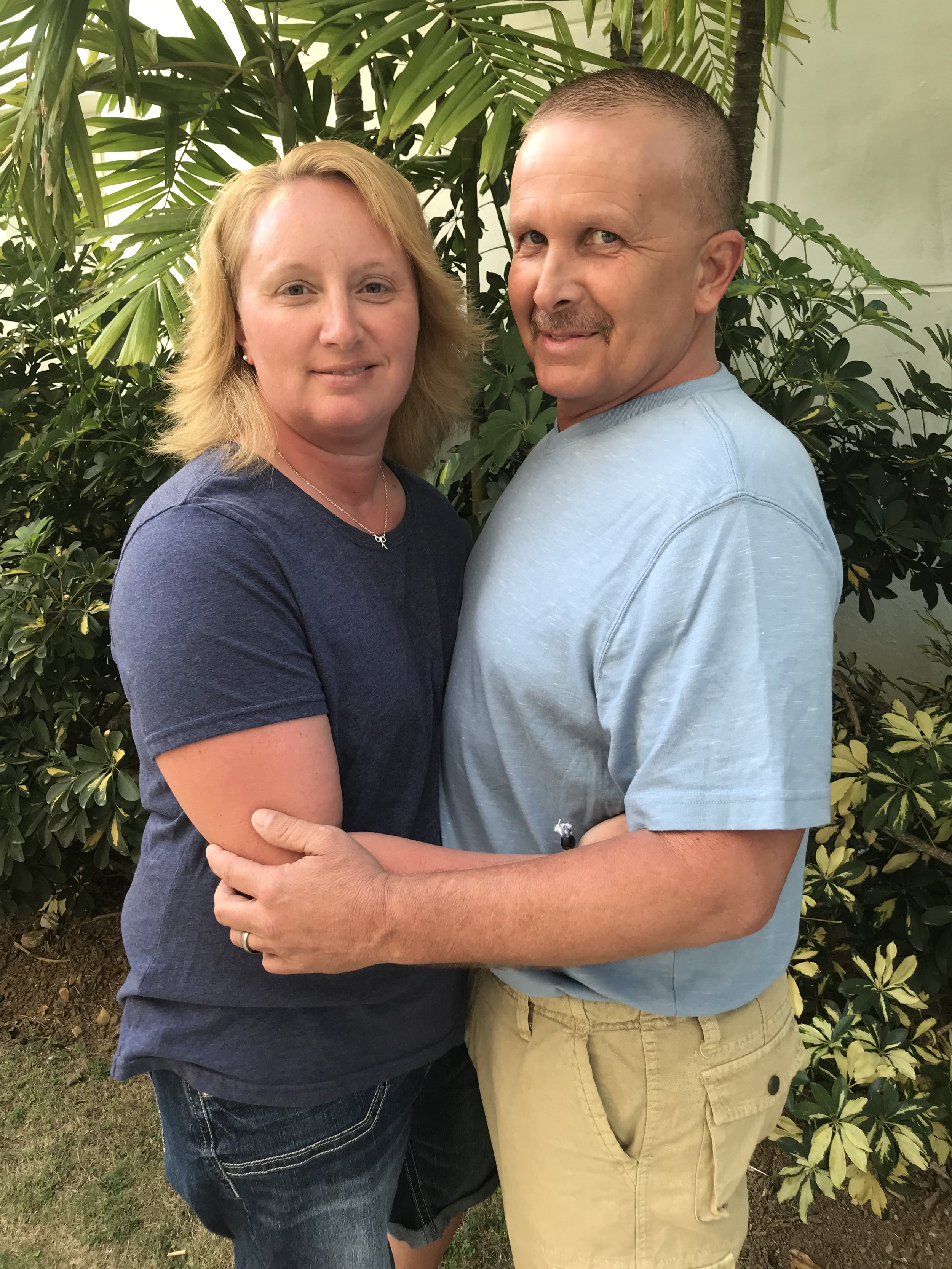 A woman with blonde hair and a man with short dark hair and a mustache holding each other in front of green tropical plants.
