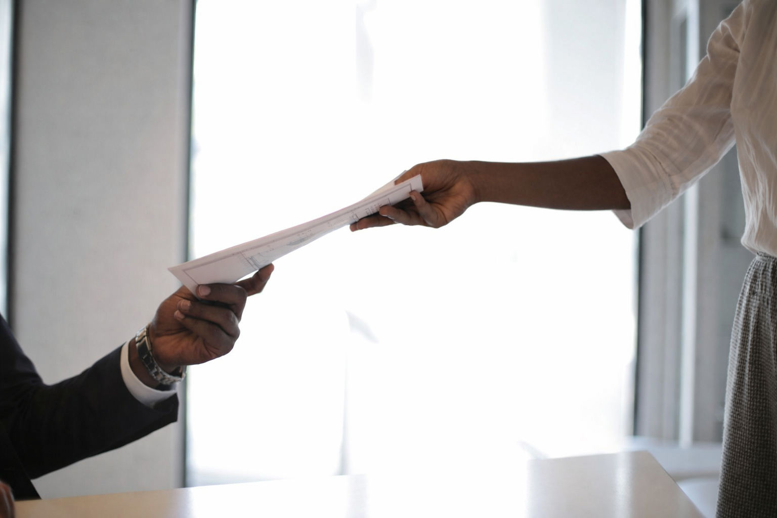 Two people exchanging a document in an office setting with bright backlighting and large windows.
