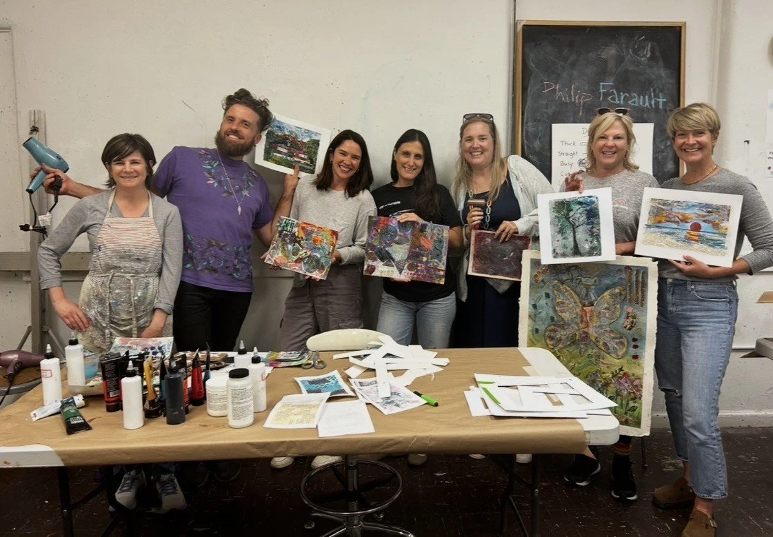 Group of seven women and one man in a classroom holding their artwork, with art supplies on the table in front of them, and a blackboard behind with the name 'Philip Farault' written on it.