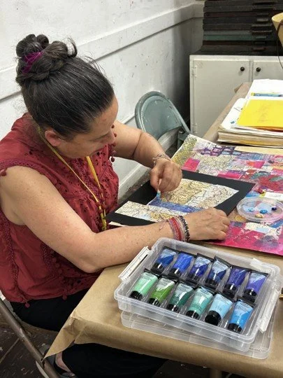 Woman in a red top working on a mixed media art project with paints and fabric, sitting at a table with art supplies and papers.
