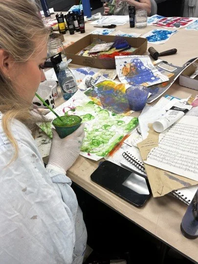 A woman working on an art project at a cluttered table with various paints, brushes, and paper.